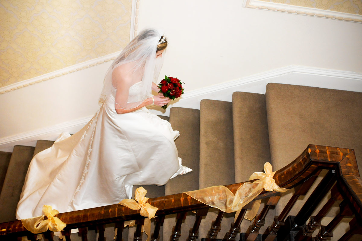 A bride wearing a white flowing wedding dress holding a bouquet of flowers climbing a grand staircase decorated with ribbons