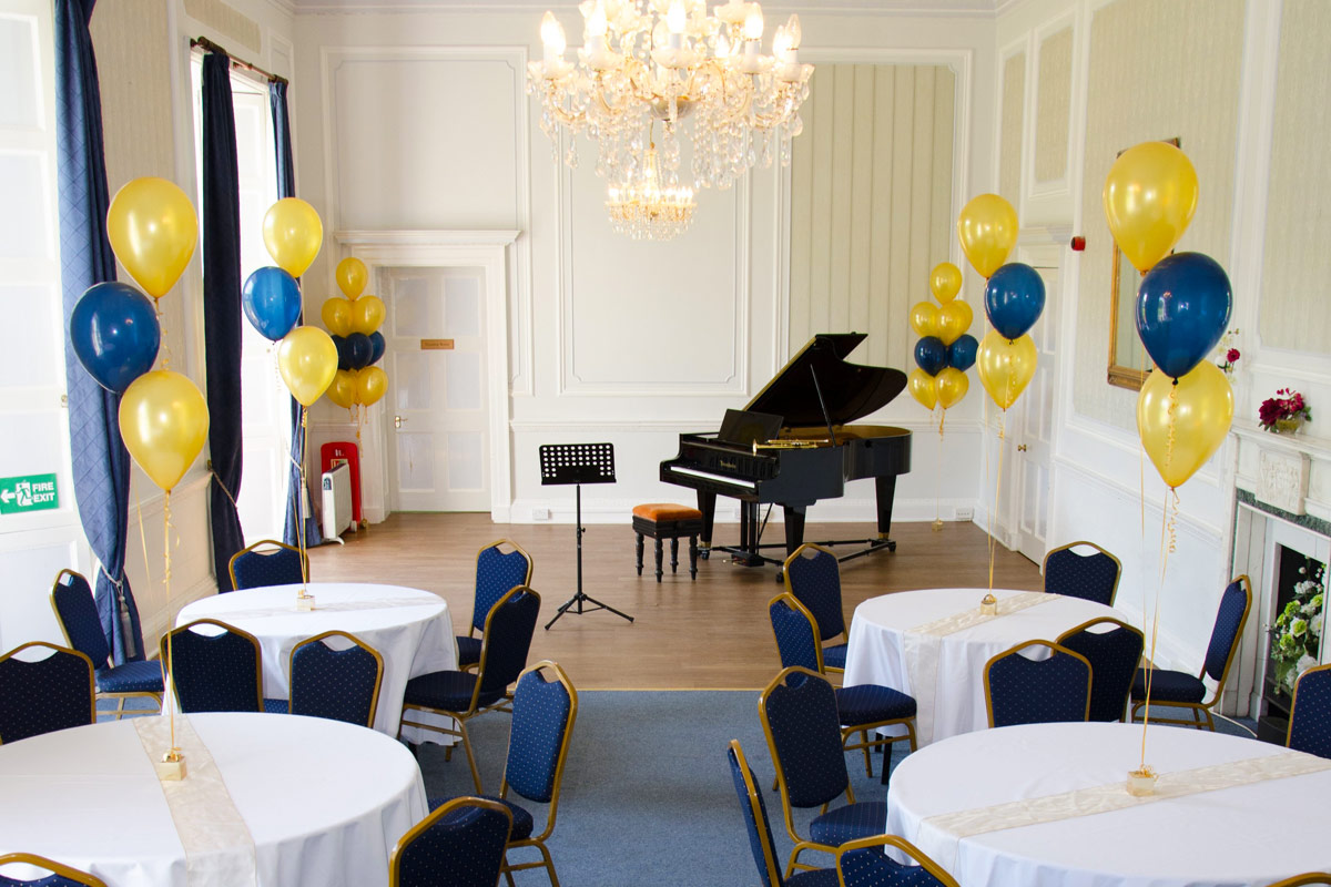 a large room set up with round tables and chairs with balloons rising from each table there is a grand piano situated at the end of the room