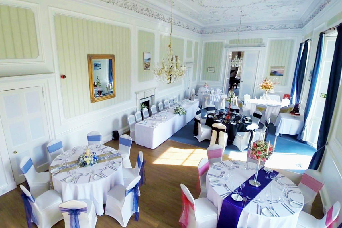 A large, elegant room set up for a wedding with multiple tables and chairs decorated with flowers and chaircovers