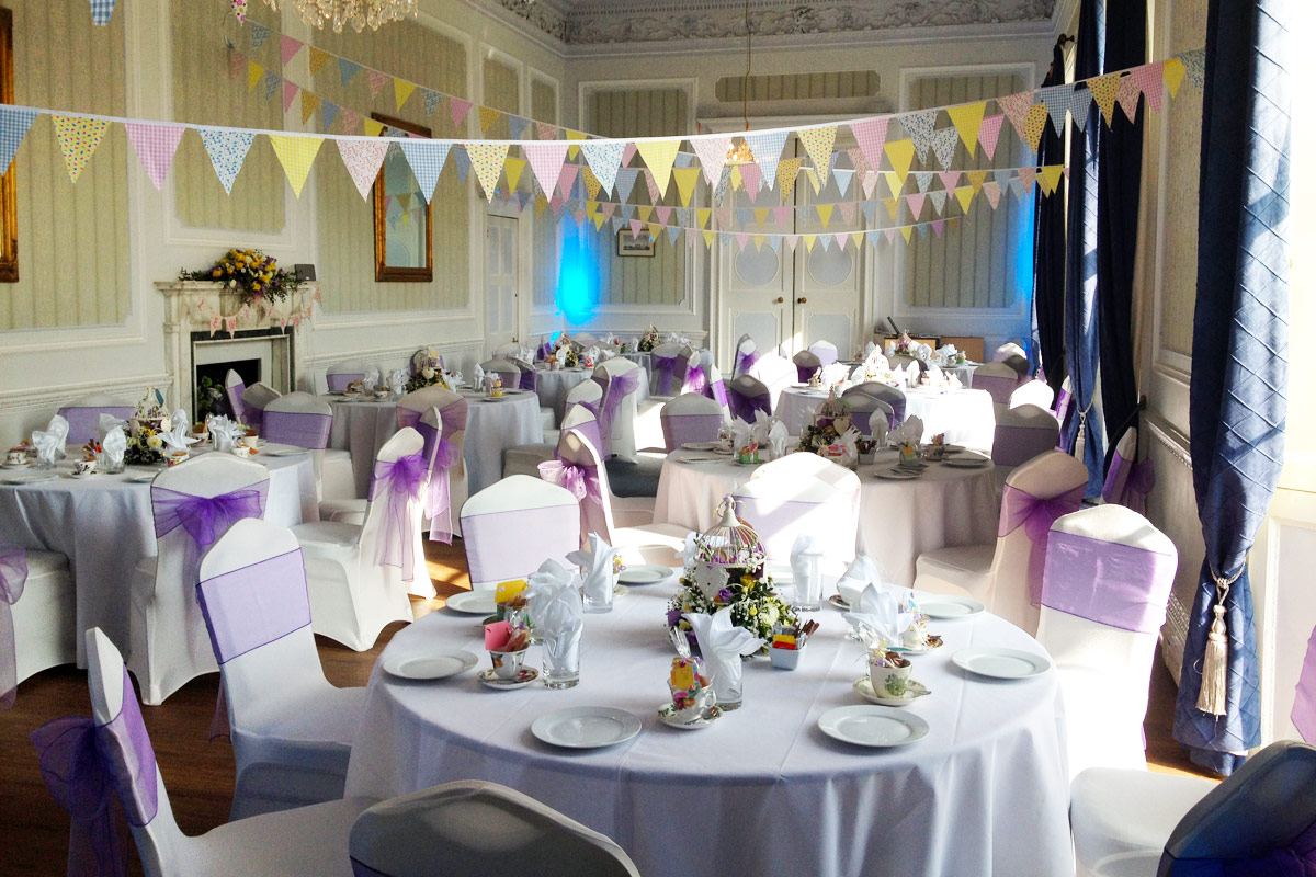 multiple elaborately decorated tables laid out with flowers and silverware. Bunting hangs from the ceiling