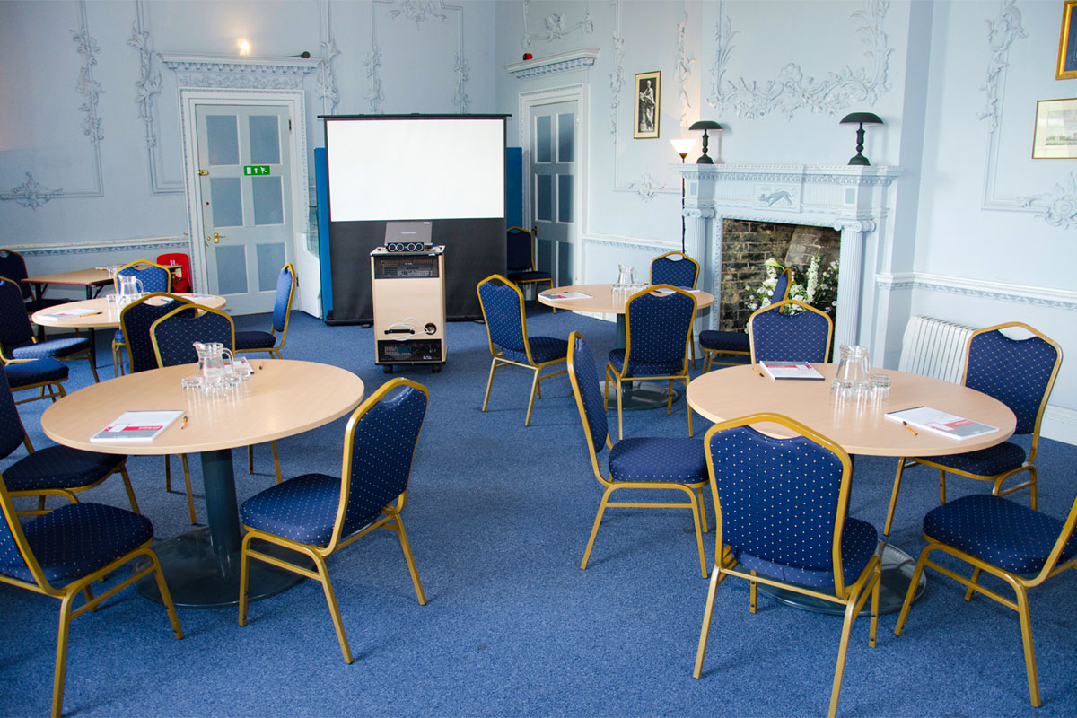 A room set up with round tables and chairs, there is a large fireplace on the back wall and a projector screen situated in the corner of the room
