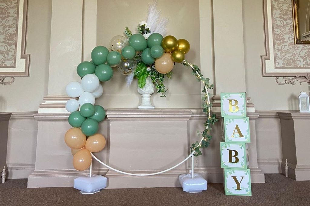 A circular white metal hoop decorated with a partial balloon arch in sage green, white, gold, and peach. To the right, a vertical stack of four light green boxes spells out "BABY" in gold lettering. The display is set against a neutral, ornate wall with a floral arrangement in a white vase centered behind the hoop.