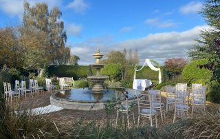 The image shows an outdoor wedding setup in a garden. In the foreground, there is a circular fountain with water, surrounded by a paved area. Around the fountain, there are white chairs arranged in rows on both sides, likely for guests. The chairs have white covers or decorations on them. In the background, there is a white arch draped with fabric. The garden is lush with greenery, including bushes and trees, and the sky is blue with some clouds. The setting appears serene and picturesque,