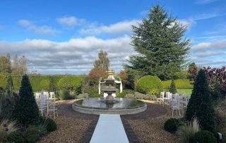 The image shows a beautifully landscaped outdoor setting prepared for a wedding. In the center, there is a circular fountain with water, surrounded by a paved area. A white aisle runner leads up to the fountain, suggesting a ceremonial path. On either side of the aisle, there are rows of white chairs arranged for guests. The background features a lush garden with various trees and shrubs, including a large evergreen tree. The sky is mostly clear with some clouds, and the overall atmosphere is serene and picturesque.