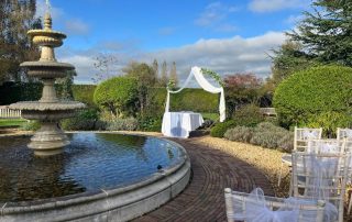 The image shows a picturesque outdoor setting prepared for a wedding ceremony. On the left side, there is a large, multi-tiered stone fountain with water gently cascading down. The fountain is surrounded by a circular pool of water. To the right of the fountain, there is a curved brick pathway leading to a small, white-draped archway decorated with greenery. Under the archway, there is a table covered with a white cloth. In the foreground on the right, there are several white chairs arranged in rows, each adorned with white tulle bows. The background features lush green hedges, trees, and a partly cloudy blue sky, creating a serene and romantic atmosphere.