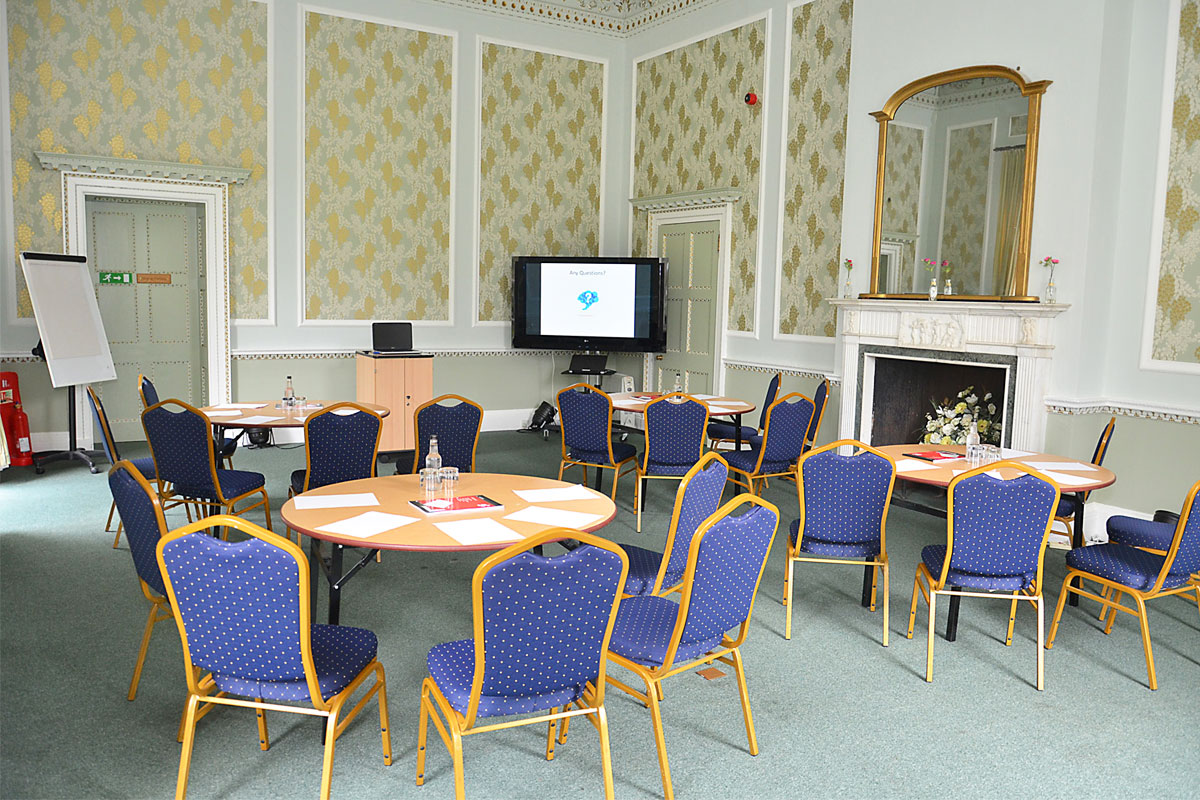A room set up with round tables and chairs, there is a large fireplace on the back wall and a large TV situated in the corner of the room