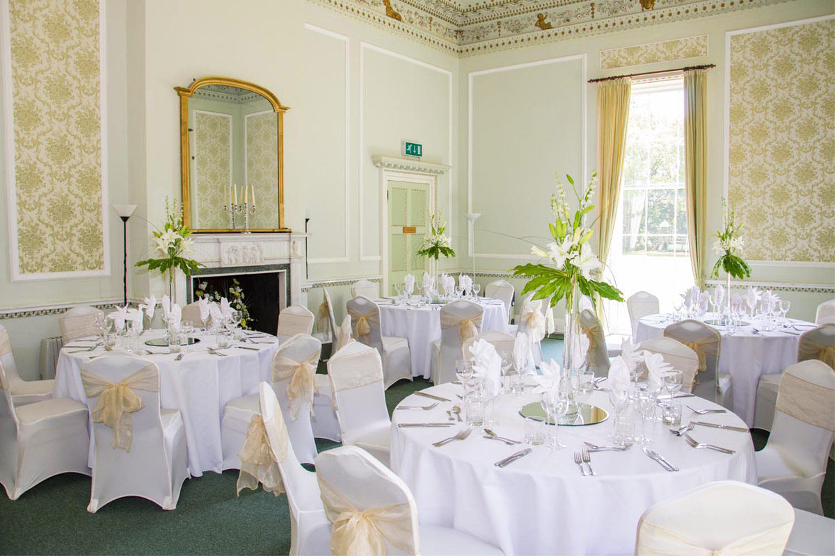 A large, elegant room set up for a wedding with multiple tables and chairs decorated with flowers and chaircovers